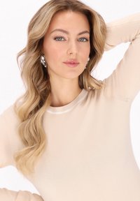 Beige, bordázott textúrájú pulóver fehér nyakkal. A modell hullámos szőke haját viseli, gyöngy fülbevalóval, az egyik kezét a feje mellett tartva pózol.