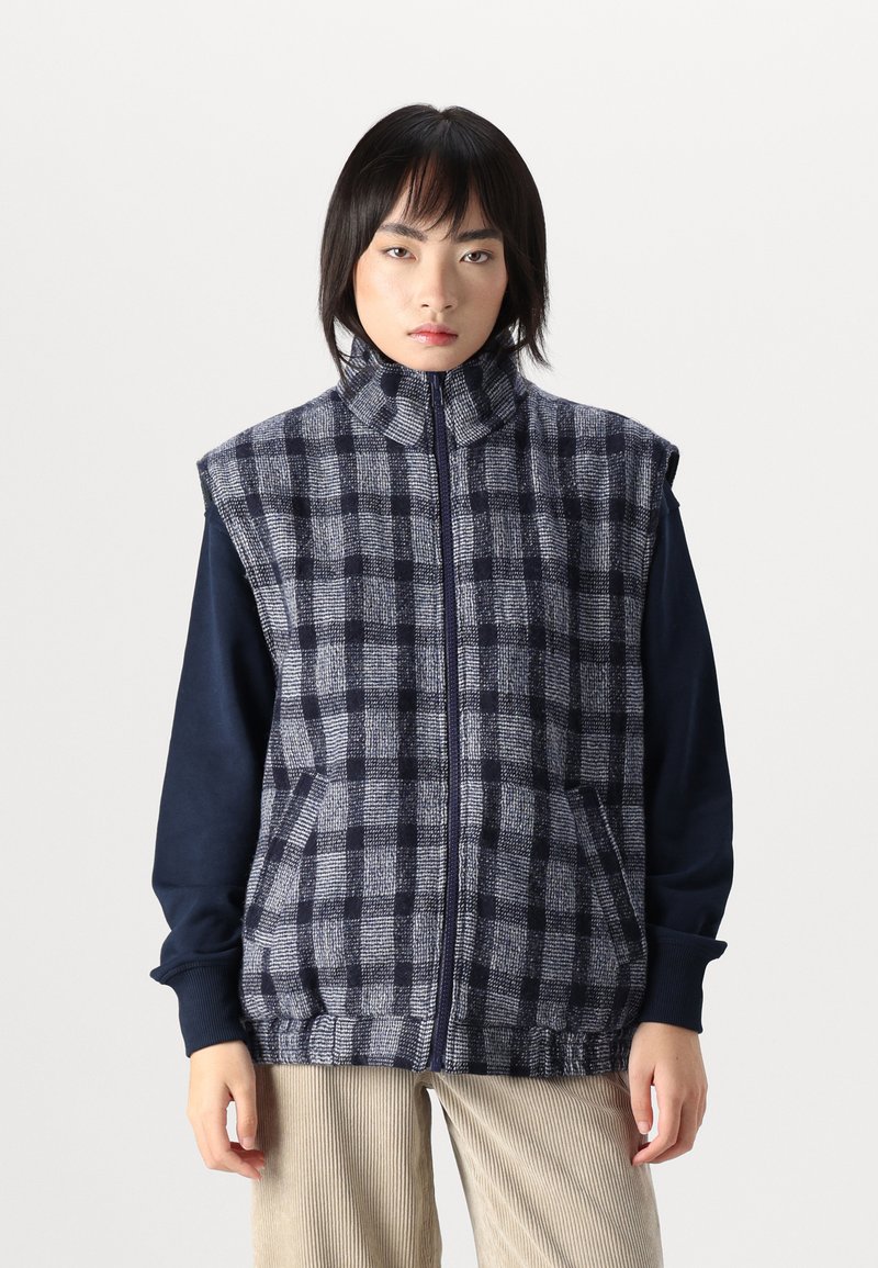 Šaha modeļu bez piedurknēm veste tumši zila un pelēka, ar augstu apkakli, ar rāvējslēdzēju un priekšējām kabatām; nēsāta pār tumšu garām piedurknēm krekli.