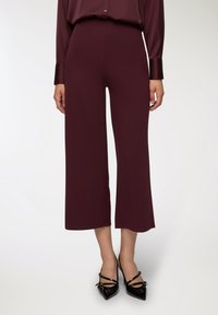 Pantaloni a gamba larga bordeaux realizzati in un tessuto morbido, caratterizzati da una vita alta e un design elegante e privo di decorazioni. Abbinati a tacchi a punta neri.