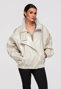 Donna che indossa una giacca di pelle color crema lucida oversize con colletto alto e orlo elastico, abbinata a pantaloni neri, in posa con le mani nelle tasche.