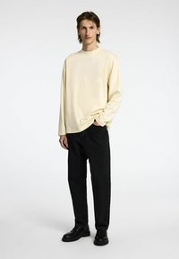 Dolgi rokav svetlo rumene srajce, kombinirane z črnimi kavbojkami in črnimi čevlji. Srajca ima okroglo ovratnico in sproščen kroj. Preprost dizajn, brez vzorcev.