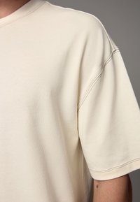 Beige cotton T-shirt with a relaxed fit, visible textured fabric, and contrasting seam details on the sleeves and neckline.