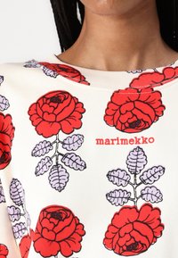 White top featuring a bold red rose pattern with purple leaves. The brand name "marimekko" is printed in red. Smooth texture and crew neck design.