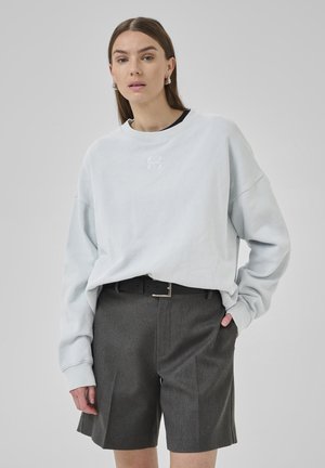 Light blue sweatshirt with a round neckline, oversized fit, and embroidered logo; paired with gray tailored shorts and a black belt.
