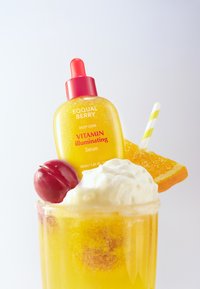 Żółta butelka serum z różową nakrętką, oznaczona napisem "Witamina Illuminating", na szczycie szklanki deserowej. Udekorowana wiśniami, plasterkiem pomarańczy i śmietaną.