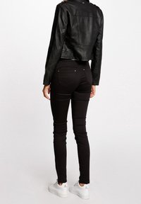 Persoon in strakke zwarte broek, korte zwarte leren jas en witte sneakers, staand met de rug naar de camera op een eentonige lichte achtergrond.