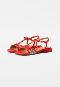 Sandali in camoscio rosso con punta aperta, design incrociato, cinturino alla caviglia e un pannello a righe bianche e nere sul lato esterno. Tacco piatto.