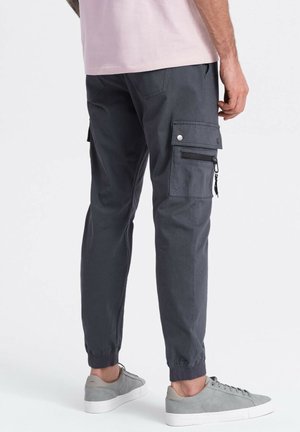 Man wearing dark grey cargo pants with side zipper pocket and light grey sneakers, shown from the back and waist down.