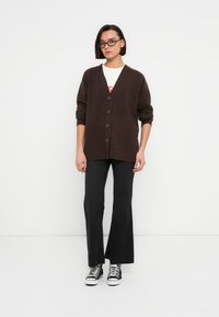 Καφέ ριμπ πλεκτή ζακέτα με V-λαιμόκοψη, πέντε κουμπιά και γυρισμένα μανίκια, συνδυασμένη με μαύρο φαρδύ παντελόνι και μαύρα-άσπρα αθλητικά παπούτσια.