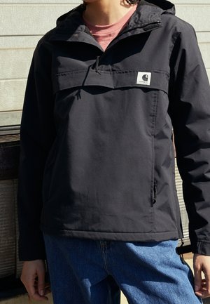 Person iført en sort Carhartt vindjakke og blå jeans, stående udendørs foran en lys væg.