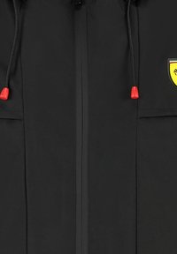 Czarna kurtka wykonana z materiału odpornego na wodę, wyposażona w zamek błyskawiczny z przodu, dwie boczne kieszenie oraz akcenty w kolorze czerwonym na sznurku. Widoczna jest także naszywka z logo.