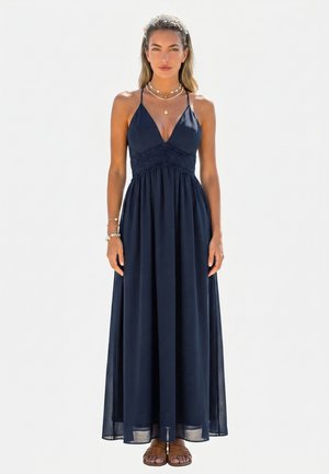 Frau trägt ein ärmelloses Maxikleid in Navyblau mit Spitzendetails, geschichtete goldene Halsketten, Armbänder, ein Blumen-Haarband und braune Sandalen.