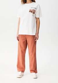 T-shirt en coton blanc avec un graphique imprimé sur la poitrine, associé à un pantalon en coton ample de couleur rouille et des baskets beiges.