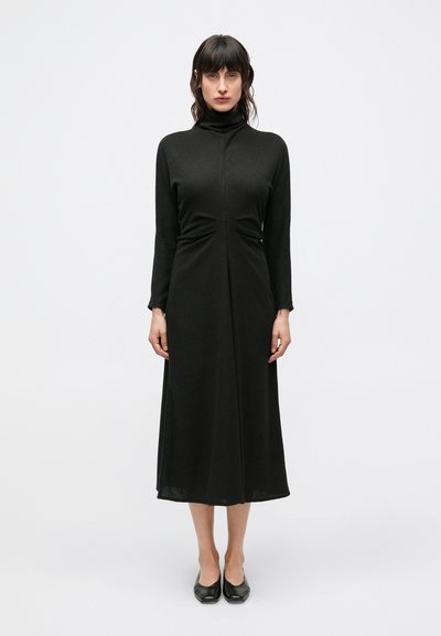 Zwarte jurk met lange mouwen, hoge hals, getailleerd lijfje, geplooide details bij de taille en een vloeiende rok, gemaakt van een gestructureerde stof.