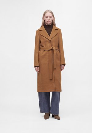 Femme portant un long manteau camel noué à la taille, un jean bleu à jambes larges, un col roulé marron et des chaussures marron à talons, debout sur un fond blanc.