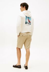 Weißer Hoodie mit einem Segelmotiv auf dem Rücken, kombiniert mit beigen Shorts. Model trägt dunkle Slipper und präsentiert ein lässiges Outfit.