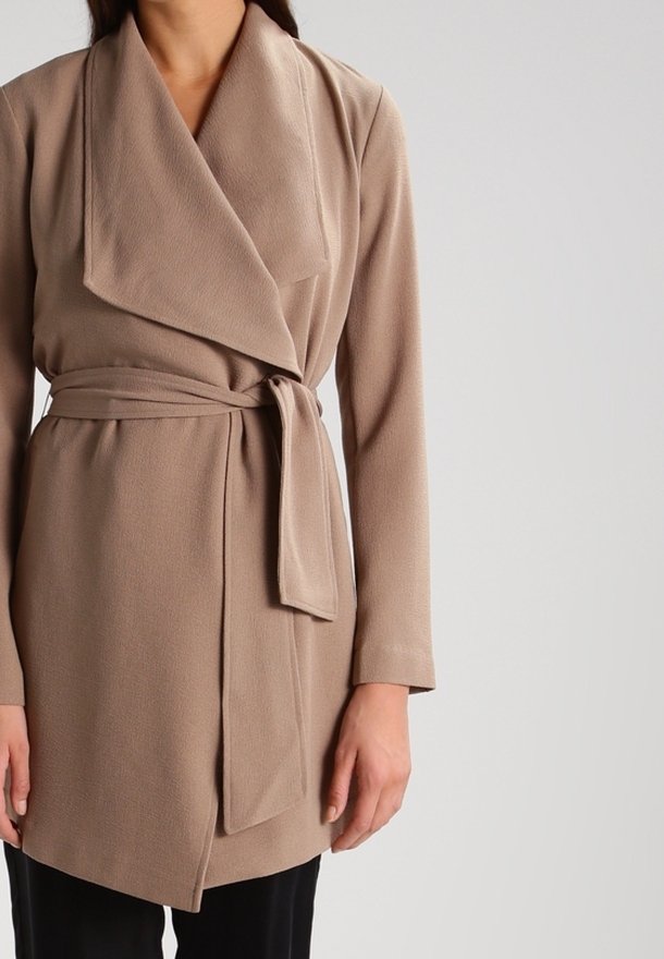 Femme portant un manteau beige ceinturé à large col et ceinture nouée, sur un pantalon noir, sur un fond uni.