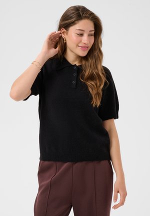Jeune femme aux longs cheveux ondulés portant un pull polo noir manches courtes et un pantalon marron, touchant son oreille et souriant doucement.