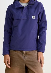 Persona que lleva una chaqueta con capucha de Carhartt púrpura con bolsillos con cremallera y pantalones marrones, de pie con las manos a los lados.