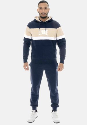 Uomo in piedi rivolto in avanti che indossa una felpa con cappuccio a righe blu scuro, beige e bianco con pantaloni sportivi blu scuro abbinati e scarpe da ginnastica bianche.