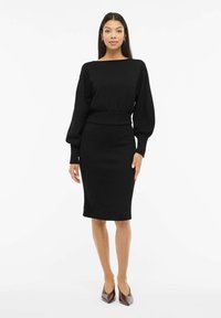 Musta, vartaloa myötäilevä mekko, jossa on pitkät, pussimaiset hihat ja leveä pääntie. Pehmeästä kankaasta valmistettu, siinä on vyötäröltä kiristetty leikkaus ja polven pituinen helma.