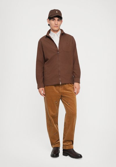 Homme portant une casquette marron, une veste zippée marron, une chemise rayée, un pantalon en velours côtelé beige avec de petites broderies, et des bottes marron foncé, se tenant contre un fond blanc.