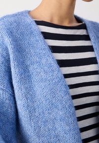 Cardigan en tricot bleu avec un tissu texturé, design à ouverture sur le devant, porté par-dessus un haut rayé noir et blanc. Gros plan sur les matériaux.