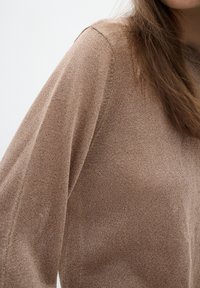 Gros plan d'une personne portant un pull en maille marron clair à manches longues, montrant la texture et le détail de la couture de l'épaule.