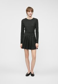 Svart topp med lange ermer og hvite prikker, kombinert med et plissé svart skjørt. Modellen har på seg svarte leiligheter. Nøytral bakgrunn.