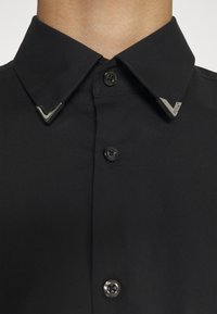 Chemise noire à boutons avec col pointu présentant des embouts de col en métal argenté en forme de V de chaque côté.