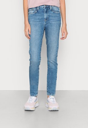 Jeans de ganga azul claro com um corte justo, apresentando bolsos frontais e um acabamento ligeiramente desbotado. Usados com ténis de desporto rosa e cinza.