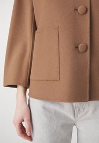 Chaqueta marrón de mezcla de lana con cuello redondeado, dos botones y un bolsillo en el pecho, combinada con pantalones vaqueros gris claro.