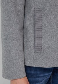 Grijze wolmix jas met een textuurstof, een ribbelende pocketdetail en een rechte snit aan de zoom. Gecombineerd met donkerblauwe jeans.