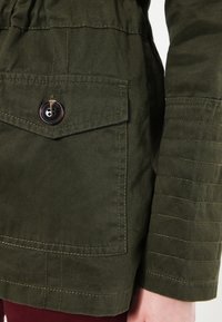 Rękaw oliwkowej kurtki z pikowanym przeszyciem i kieszenią na klapę zapinaną na jeden ciemny guzik, noszonej na burgundowych spodniach.