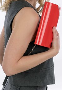 Clutch de charol rojo con una forma rectangular elegante, sostenido contra una blusa sin mangas gris, con una delgada correa de cadena plateada.
