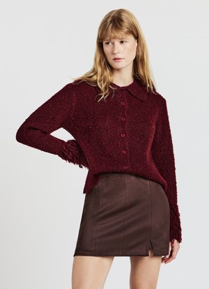 Bordeauxroter, strukturierter Strickpullover mit Kragen und Knopfverschluss, kombiniert mit einem braunen Rock aus Kunstleder mit Seitenschlitz.