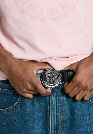 Manos de piel oscura ajustando un cinturón negro con hebilla circular plateada con un diseño detallado, usado con vaqueros azules y una camisa rosa.