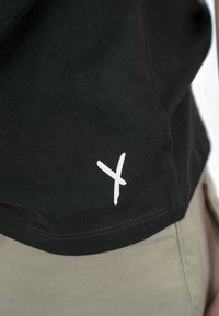 Juodas medvilninis marškinėlis su trumpomis rankovėmis, su baltu logotipo spaudiniu apatiniame kairiajame kampe. Lygus audinys, įprasta siluetas.