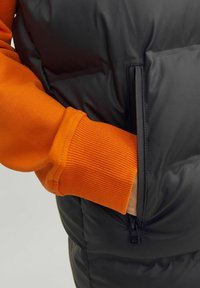 Polstret vest med et sort yderlag og en sidelomme med lynlås, parret med et orange ribbet manchet, der ses ved ærmet.