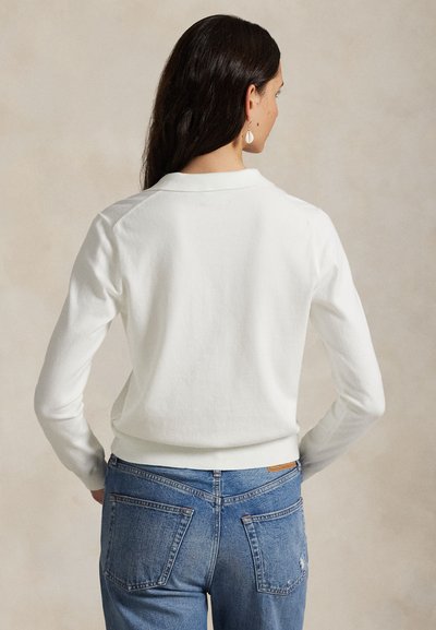 Bela majica z dolgimi rokavi, rebrastimi manšetami in okroglim izrezom, kombinirana z modrimi kavbojkami z zadnjimi žepi in sproščenim krojem.