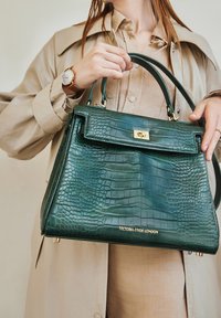 Zielona torebka w kroku krokodyla z złotymi elementami i zapięciem na klamrę. Posiada podwójne uchwyty i usztywnioną konstrukcję. Widoczna nazwa marki.