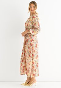 Blumen-Maxi-Kleid mit einer cremefarbenen Basis und roten sowie grünen Mustern, langen Puffärmeln und taillierter Taille. Kombiniert mit goldenen Slingback-Absätzen.