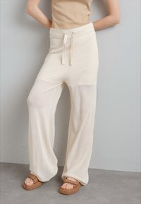 Pantalon en mesh léger de couleur crème avec une coupe décontractée, une taille élastique et un cordon de serrage. Associé à des sandales brunes chunky.