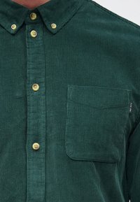 Camicia verde in velluto a coste con colletto con bottoni, bottoni gialli e taschino sul petto con un'etichetta logo discreta. Tessuto strutturato con coste verticali.