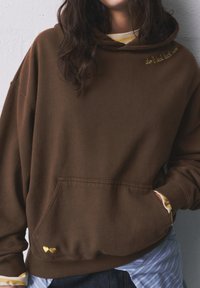 Hnedá kapucňová mikina s predným vreckom; obsahuje žltý vyšívaný text a malé srdcové akcenty. Nosená na pruhovanej košeli.