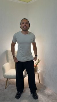 Uomo in piedi in una stanza che indossa una maglietta grigio chiaro, pantaloni neri e scarpe nere, con una sedia e un tavolino con un vaso dietro di lui.