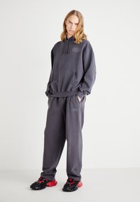 Szürke, oversized kapucnis pulóver elülső zsebbel és mellkasi logóval, hozzá illő melegítőnadrággal, amelyen egy kis logó található. Fekete és piros sportcipők.