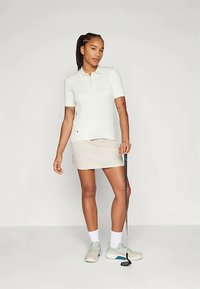 Λευκό αθλητικό πουκάμισο με ριμπ και ανοιχτό μπεζ φούστα. Το μοντέλο κρατά μια σκανδάλη γκολφ, φορώντας γκρι και μπλε αθλητικά παπούτσια με λευκές κάλτσες.