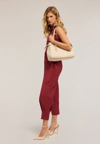 Beżowa torebka o zaokrąglonym kształcie, gładkiej fakturze i kontrastowym czarnym wykończeniu; noszona z bordowym kombinezonem i beżowymi szpilkami.