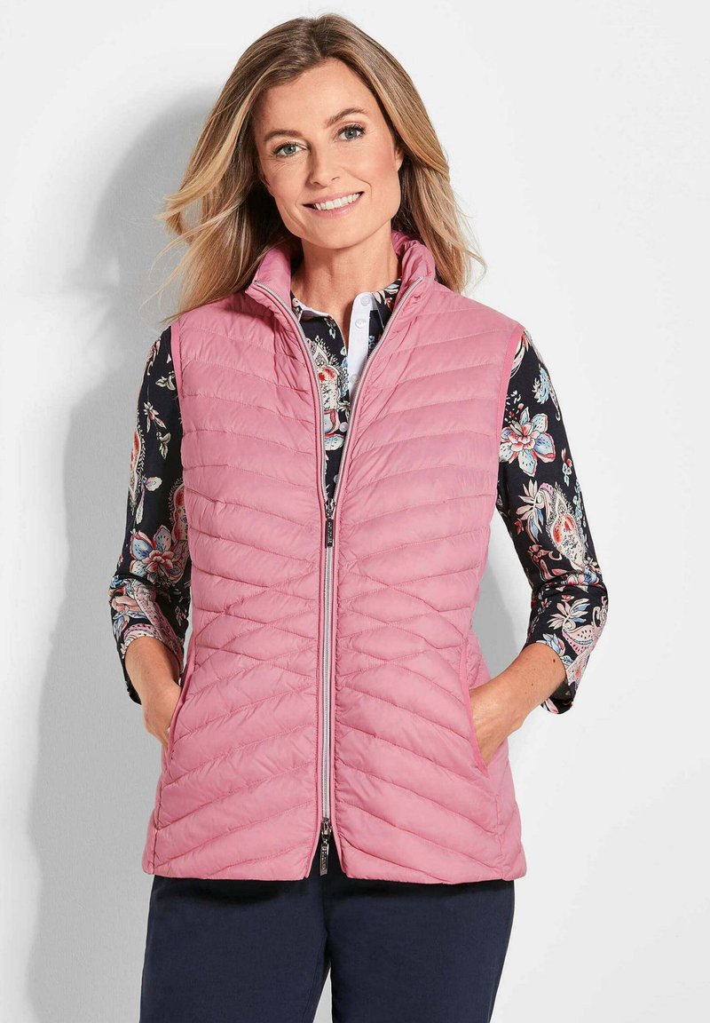 Gepolsterte rosa Weste mit hohem Kragen, Frontreißverschluss und Seitentaschen, über einem langärmligen, dunkelblumigen Hemd getragen.